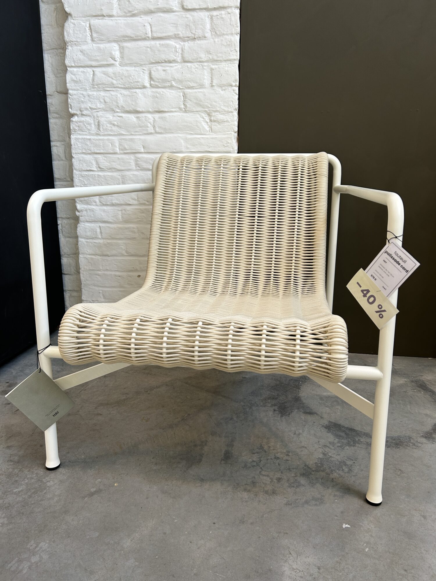 Une chaise Expo - Fauteuil bas Palissade Cord à armature métallique blanche, avec une assise et un dossier en rotin tressé, est exposée à l'intérieur, avec une étiquette indiquant une réduction de 40 %.