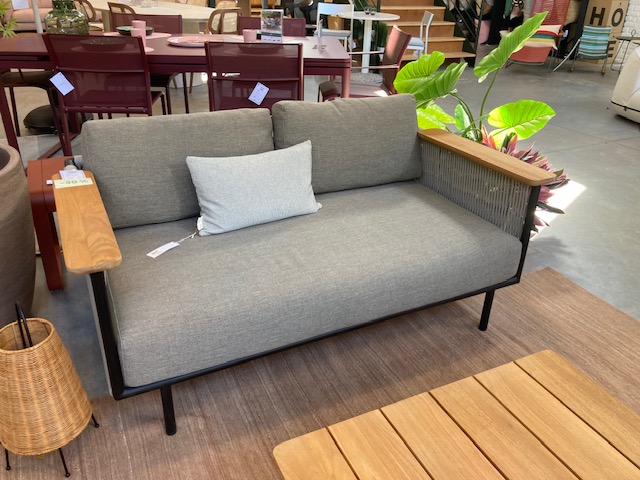 Un Expo - Canapé de jardin Corbey avec des coussins gris et des accoudoirs en bois est placé sur un tapis marron à côté d'une table en bois et d'une plante en pot dans une salle d'exposition.