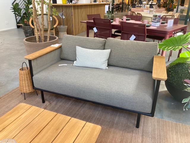 Expo moderne grise à deux places - Canapé de jardin Corbey avec accoudoirs en bois et un seul coussin, présenté sur un tapis marron dans une salle d'exposition aux côtés de tables, de chaises et de plantes.