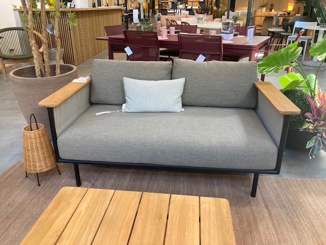Un canapé deux places gris moderne avec des accoudoirs en bois et un coussin léger, similaire à l'Expo - Canapé de jardin Corbey, est posé sur un tapis marron à côté d'une lampe tressée et d'une table basse en bois dans une salle d'exposition.