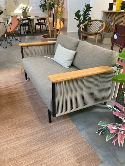Une causeuse grise moderne, Expo - Canapé de jardin Corbey, avec des accoudoirs en bois et des détails latéraux en corde, présentée sur un tapis marron clair dans une salle d'exposition contemporaine.