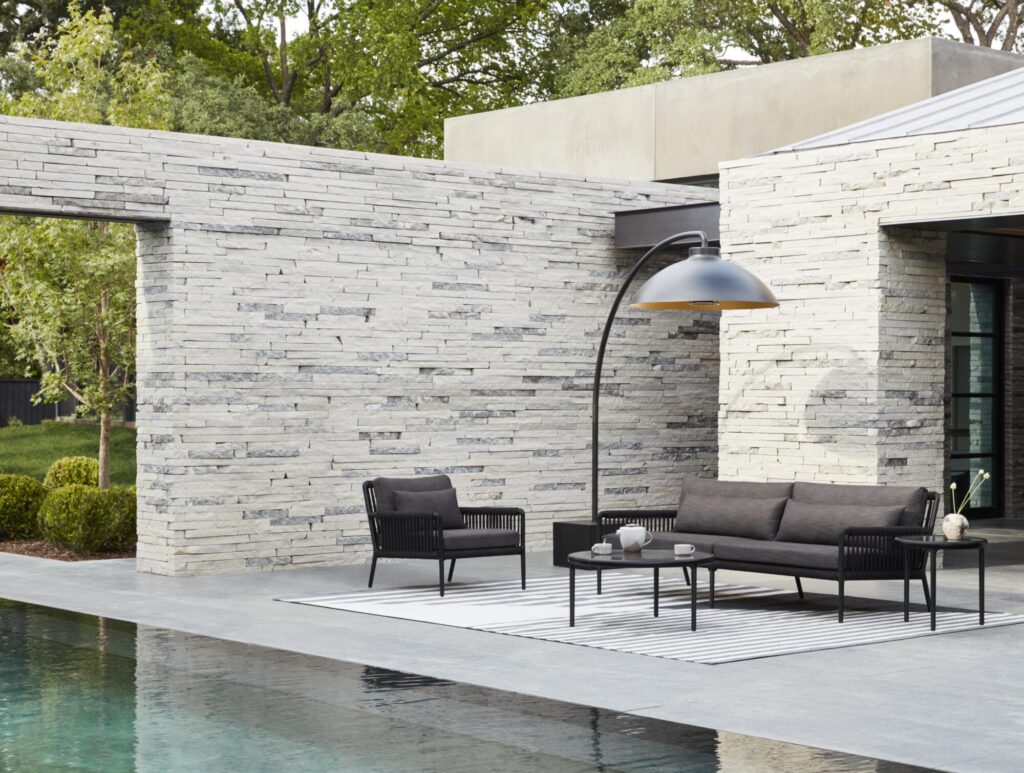 Patio extérieur moderne avec des murs en pierre, un canapé, deux chaises, une table basse et une grande lampe DOME FS incurvée qui projette des ombres de minuit sur le tapis rayé près de la piscine, avec des arbres et des touches de neige à l'arrière-plan.