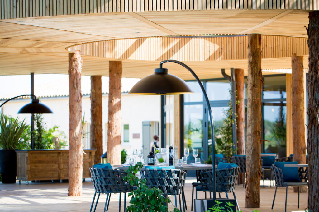 Patio du restaurant en plein air avec plafond en bois, colonnes en tronc d'arbre, tables et chaises modernes garnies de verres et d'une grande lampe DOME FS en surplomb - parfait pour profiter d'un dîner de minuit sous les étoiles.