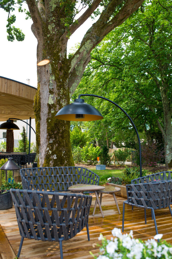 Terrasse extérieure avec deux chaises noires tressées de la collection DOME FS, une table en bois et une grande lampe sous un arbre, installée sur une terrasse en bois entourée de verdure luxuriante pour des réunions douillettes à minuit.