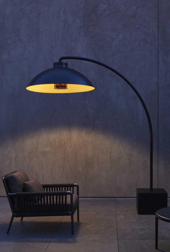 Un lampadaire moderne DOME FS noir s'arque au-dessus d'une chaise d'extérieur coussinée, jetant une lueur chaude de minuit sur un patio en béton avec un mur gris texturé en arrière-plan.