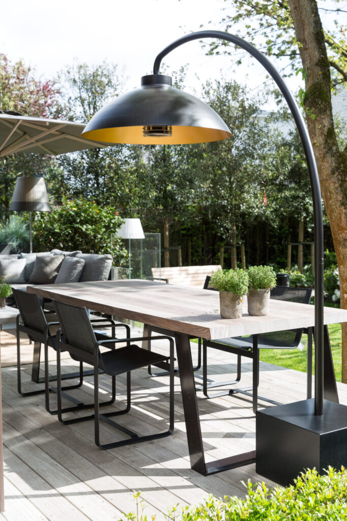 Salle à manger moderne en plein air avec une table en bois, des chaises en métal noir, deux plantes en pot et une grande lampe à arc noire sur une terrasse en bois entourée de verdure - parfaite pour les réunions de minuit sous les étoiles ou pour admirer la douceur de la neige.