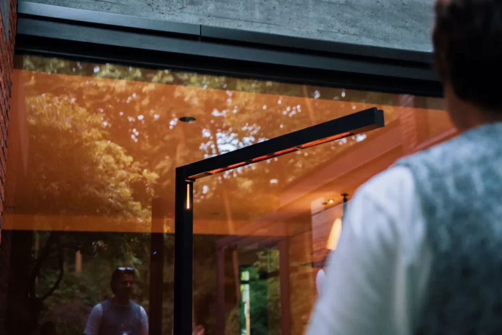 Une personne se tient près d'une porte vitrée pivotante qui reflète des arbres, une autre personne et le subtil marquage "TWIG FS - Midnight (230V, EU)" dans le verre sous la douce lumière de minuit.