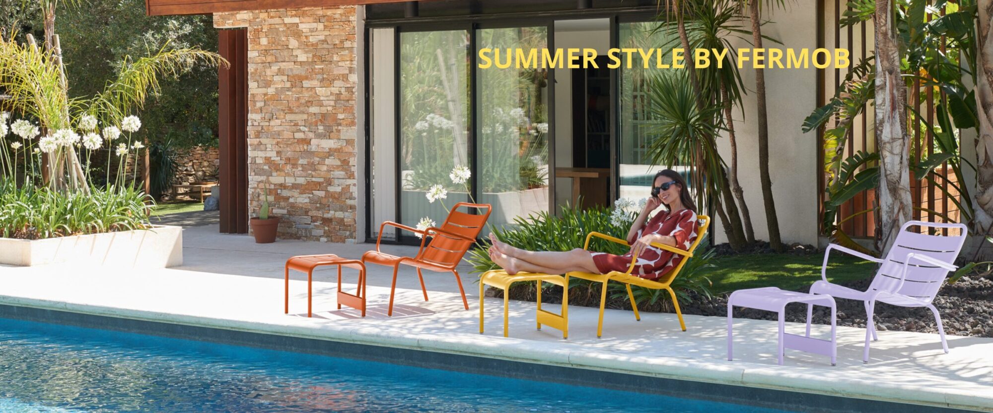 Une femme en robe rouge se détend sur une chaise jaune au bord d'une piscine, entourée de chaises d'extérieur colorées et de verdure. Le texte indique "SUMMER STYLE BY FERMOB. Soldes BE-FR 2025.