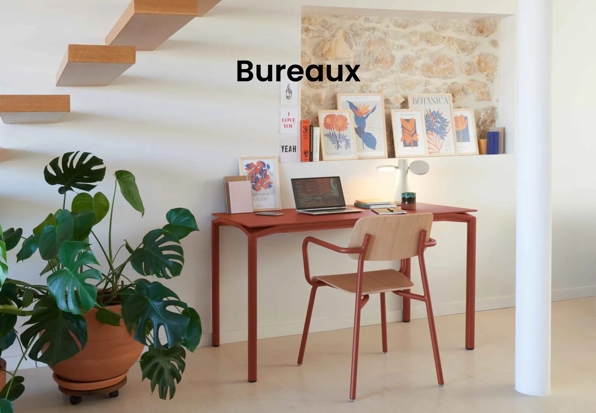 Le bureau minimaliste de Quatuor Design est composé d'un bureau et d'une chaise en bois, d'un ordinateur portable, d'une plante en pot, d'une lampe de bureau et d'œuvres d'art encadrées sur une étagère contre un mur blanc et en pierre - une inspiration parfaite pour le design d'un magasin de meubles.