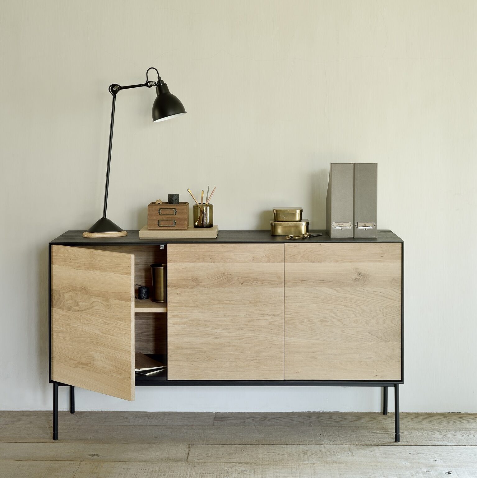 Un buffet en bois Ethnicraft pas cher avec des pieds noirs et un plateau noir contient une lampe de bureau, des fournitures de bureau et des dossiers ; une porte de l'armoire est légèrement ouverte, révélant des étagères à l'intérieur.