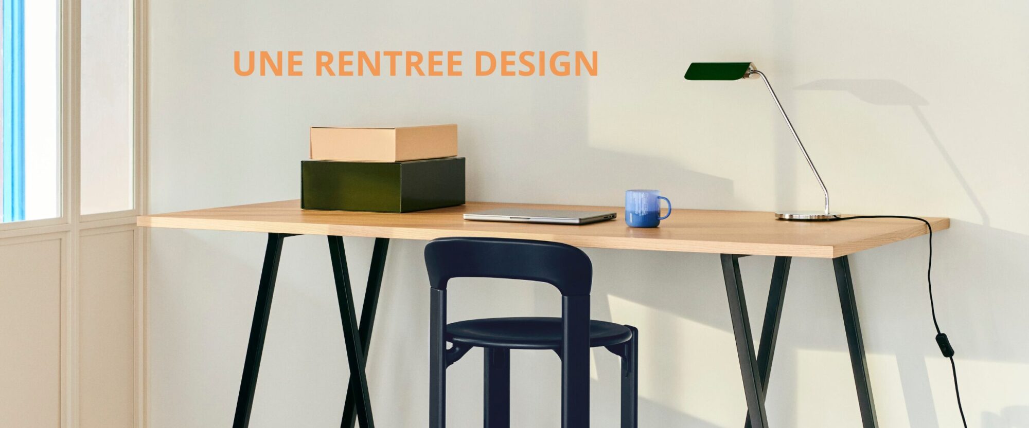 Un bureau minimaliste avec deux boîtes, un ordinateur portable, un mug bleu et une lampe de bureau moderne ; le texte sur le mur indique "UNE RENTREE DESIGN". Parfait pour votre espace de travail BE-NL-FR ou comme source d'inspiration pour votre curseur principal.