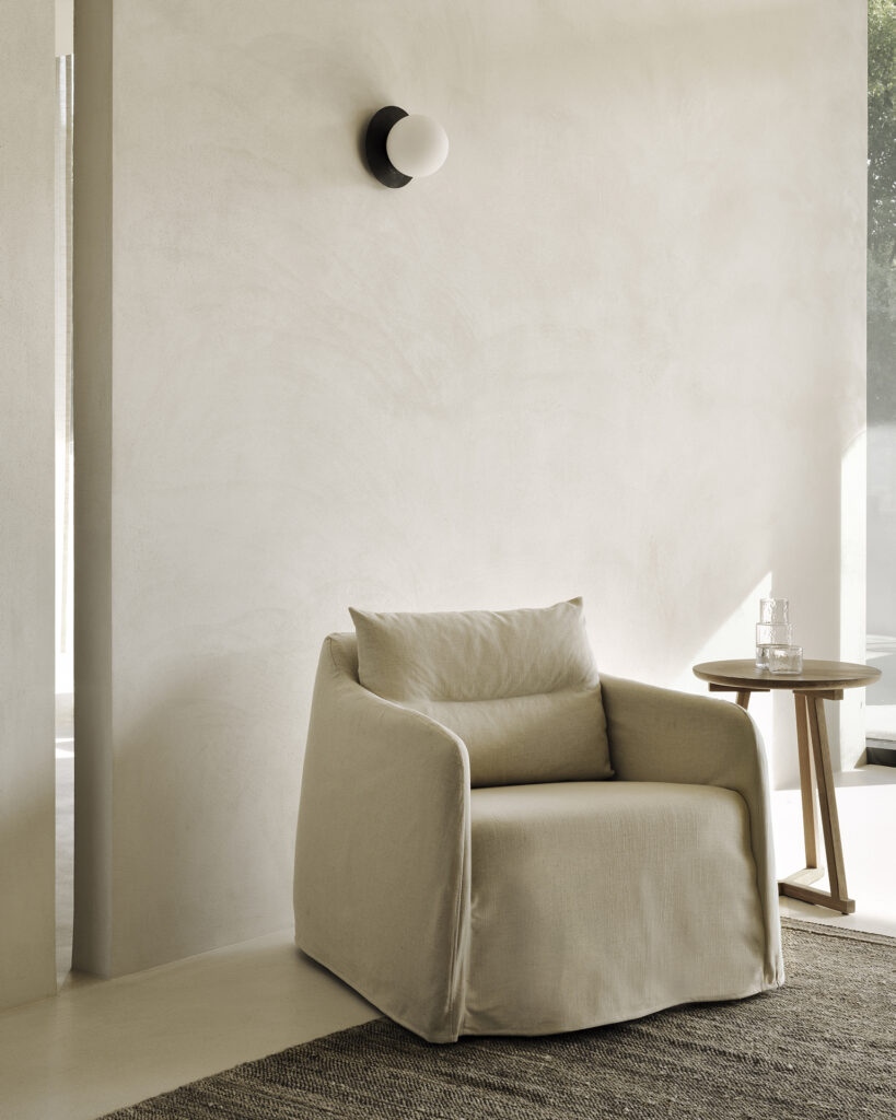 Une chaise longue en os avec un coussin est posée à côté d'une petite table d'appoint en bois contenant un verre et une carafe, sur un mur clair et texturé avec une applique murale ronde.