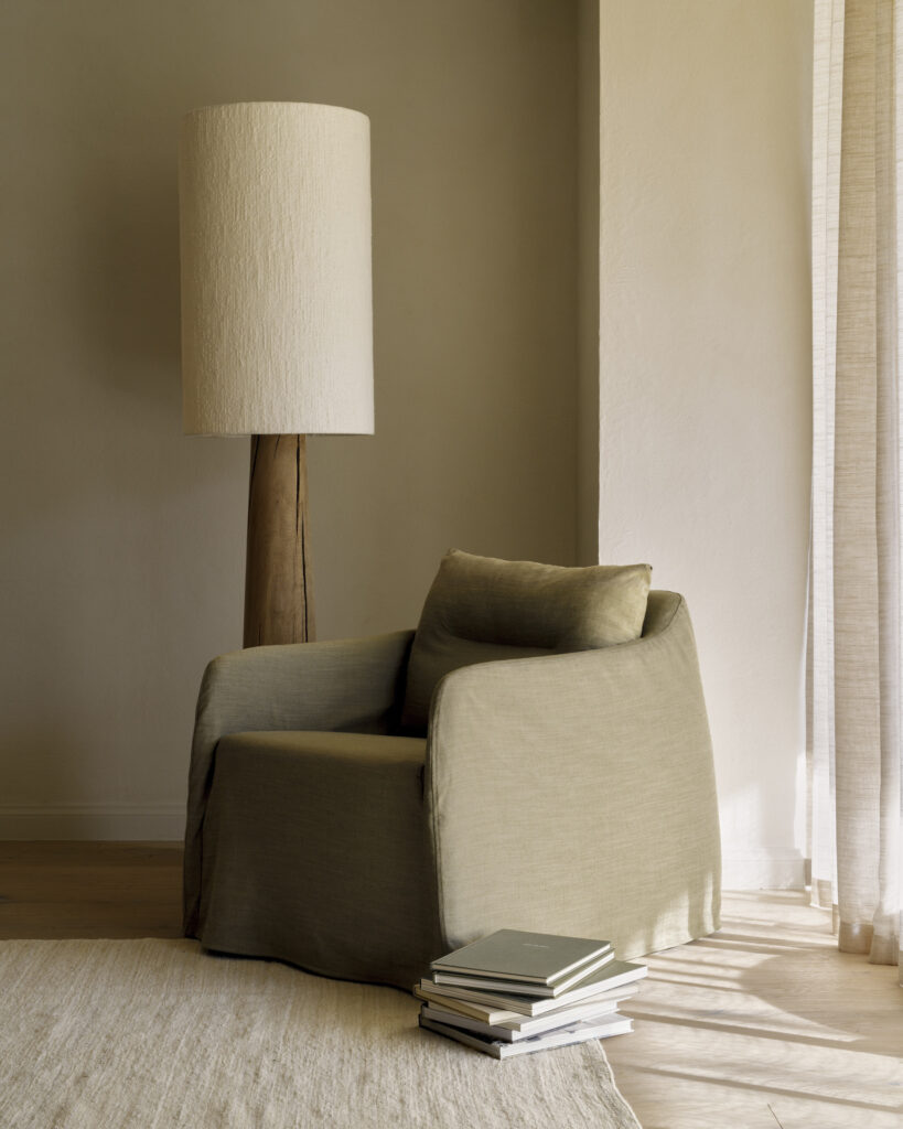 Une chaise longue Weave beige avec un coussin est assise à côté d'un grand lampadaire avec un abat-jour texturé. Plusieurs livres sont empilés sur le sol à côté de la chaise, sur un tapis clair.