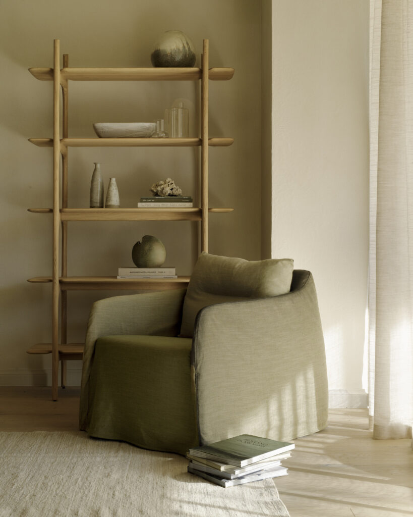 Une chaise longue en tissu vert est posée à côté d'une étagère en bois sur laquelle sont disposés des vases et des objets de décoration. Une pile de livres est posée sur le sol à proximité dans une pièce minimaliste éclairée par le soleil.