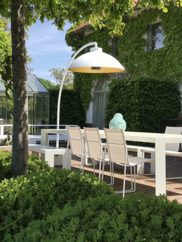 Salle à manger contemporaine en plein air avec une table et des chaises blanches sous le Lampadaire chauffant DOME FS, avec de la verdure vibrante et un bâtiment recouvert de lierre à l'arrière-plan.