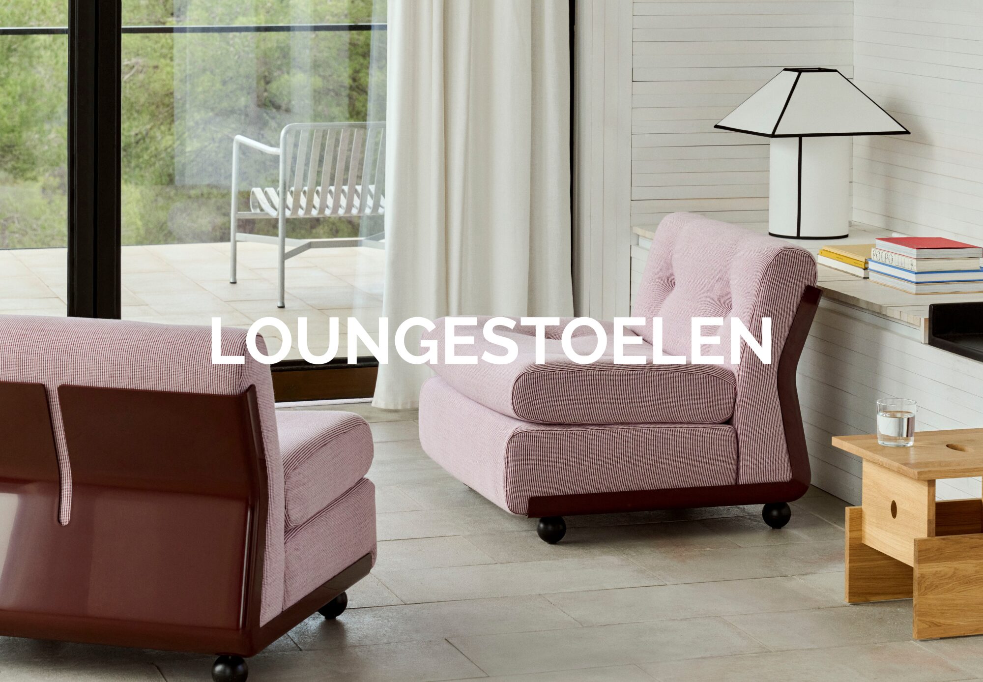 Deux chaises longues modernes mauves de Quatuor Design avec des dossiers brillants dans un salon lumineux, près d'une table d'appoint en bois, de livres empilés, d'une lampe et de grandes fenêtres. Le texte "LOUNGESTOELEN" est superposé. Les meubles design sont omniprésents.