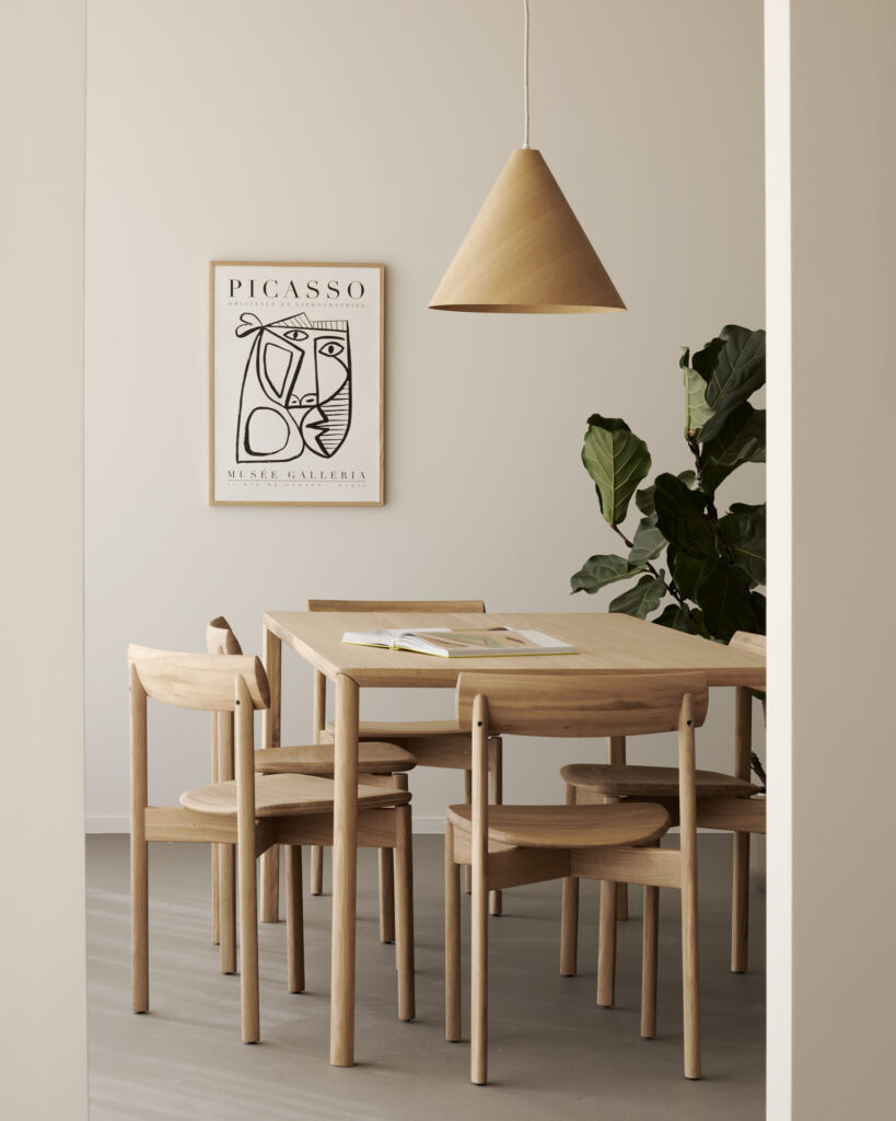 Une salle à manger minimaliste avec une table en bois clair, des chaises Risu, une lampe suspendue, une œuvre d'art de Picasso au mur et une grande plante en pot dans le coin.