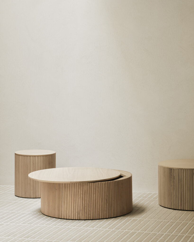 Trois tables rondes Roller Max basse en bois clair et à rainures verticales sont disposées sur un tapis quadrillé de couleur crème contre un mur beige uni.