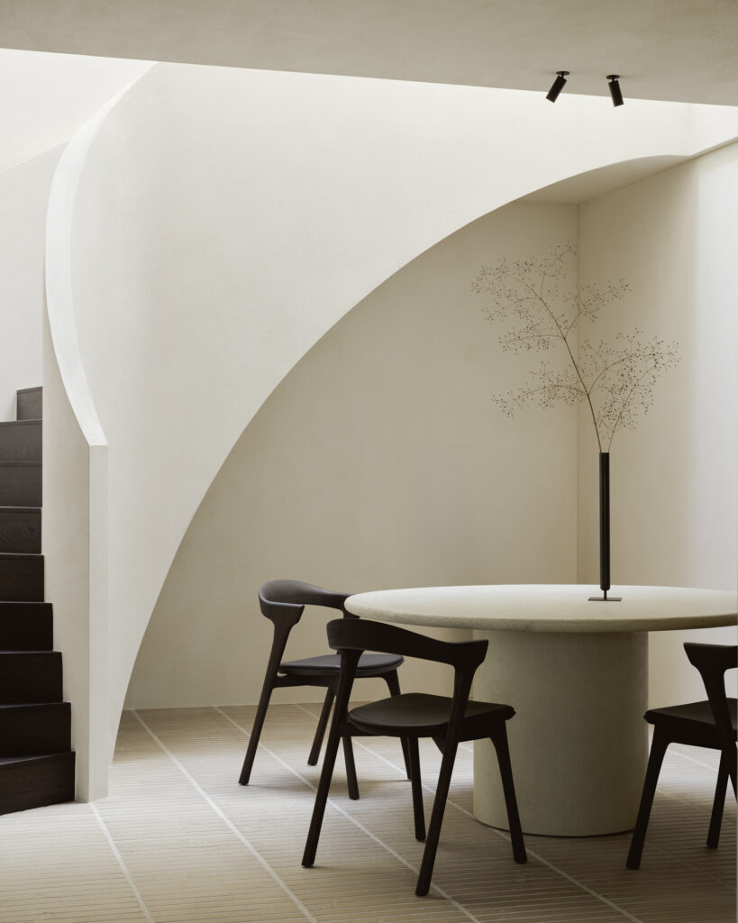 Salle à manger minimaliste avec une table ronde aux éléments raffinés, trois chaises sombres, un grand vase à la branche délicate et un escalier incurvé dans une pièce aux tons neutres baignée de lumière naturelle.