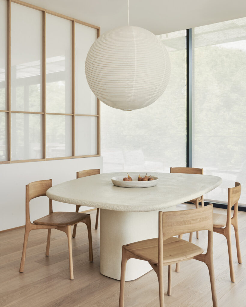Une salle à manger moderne avec une table ronde blanche aux lignes épurées, quatre chaises en bois, une grande lanterne en papier et un bol de poires, le tout entouré de grandes fenêtres et d'un décor minimaliste.