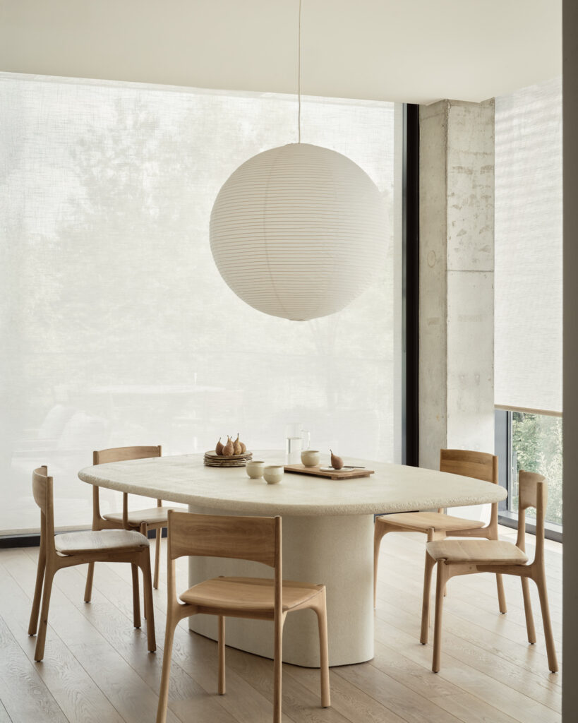 Une salle à manger minimale avec une table oblongue en bois clair, quatre chaises assorties, une grande lanterne ronde en papier et de grandes fenêtres avec des stores transparents met en valeur les éléments simples de la table et le design épuré.