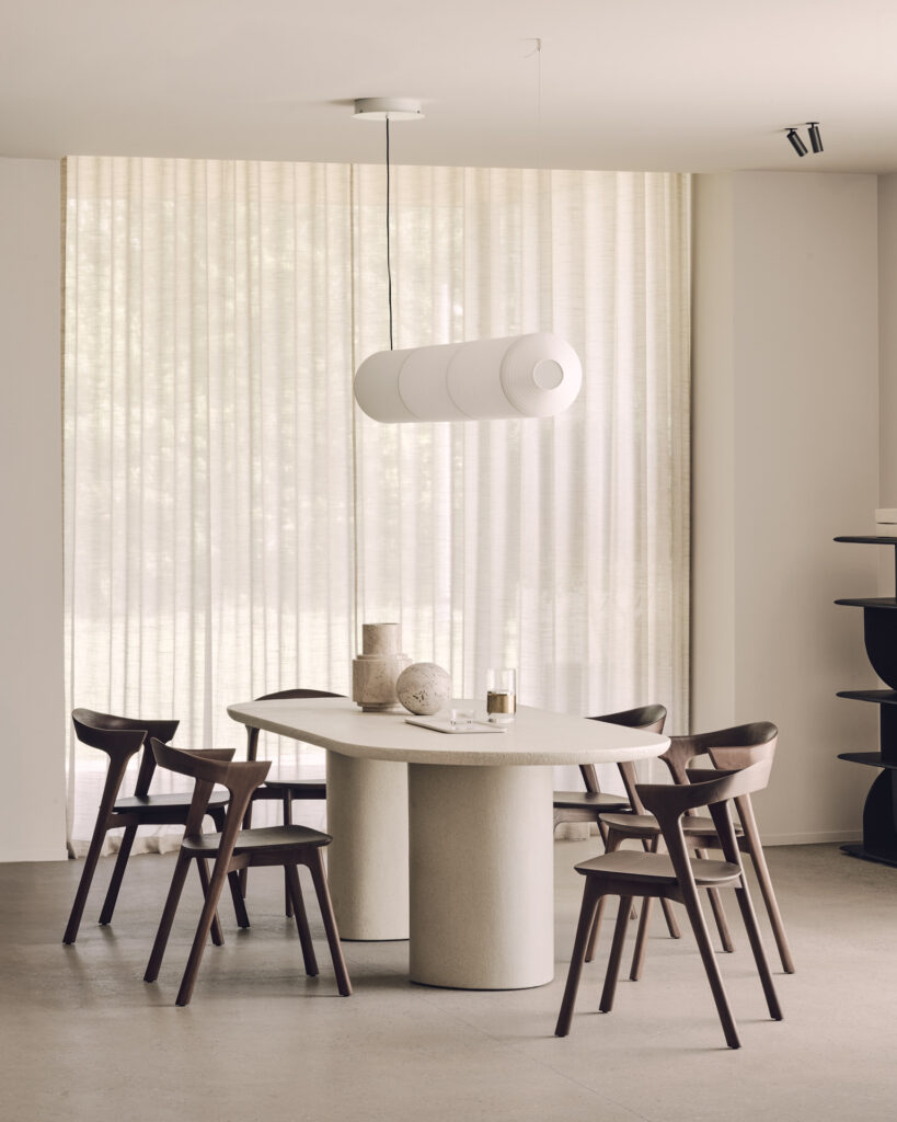 La salle à manger moderne se compose d'une table oblongue, entourée de quatre chaises en bois, d'une lampe suspendue sculpturale et de rideaux transparents allant du sol au plafond, qui laissent entrer la lumière naturelle.