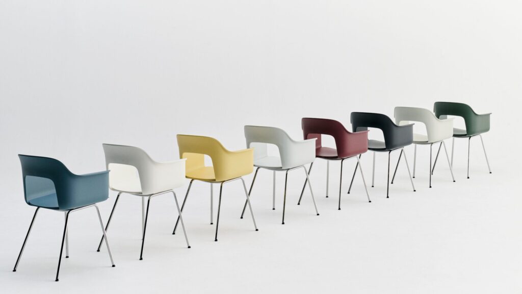 Une rangée de neuf chaises Layout avec accoudoirs et pieds en métal, chacune de couleur différente, disposées en diagonale sur un fond blanc.