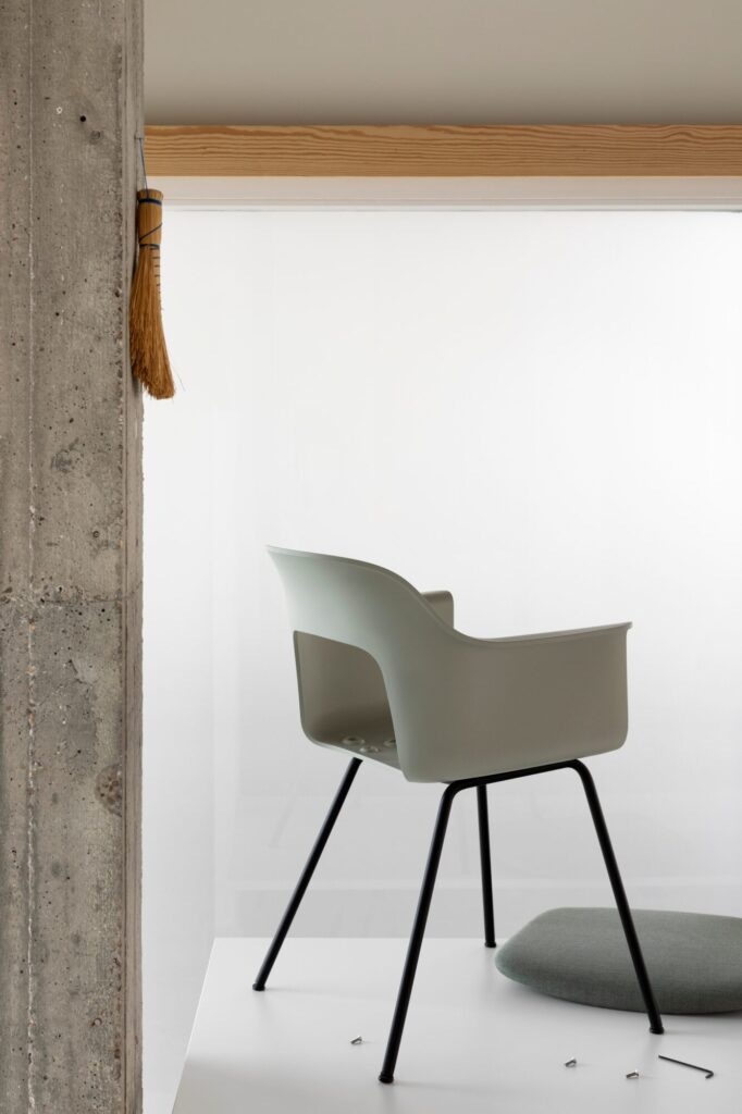 La chaise Layout, dotée d'une assise moderne en plastique gris clair et de pieds noirs, est présentée sur une plate-forme blanche à côté d'un coussin rond gris et d'un mur en béton, avec un petit balai suspendu à proximité.