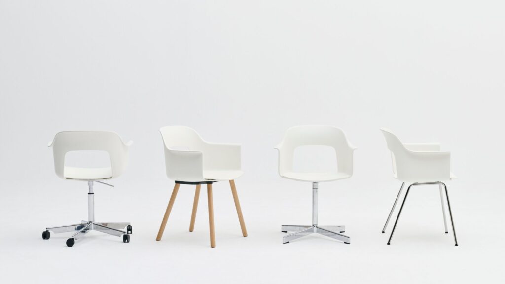 Quatre chaises blanches modernes "Layout" avec accoudoirs - chacune avec un piétement à roulettes, en bois, en métal pivotant et en métal - sont disposées en rangée sur un fond blanc uni.