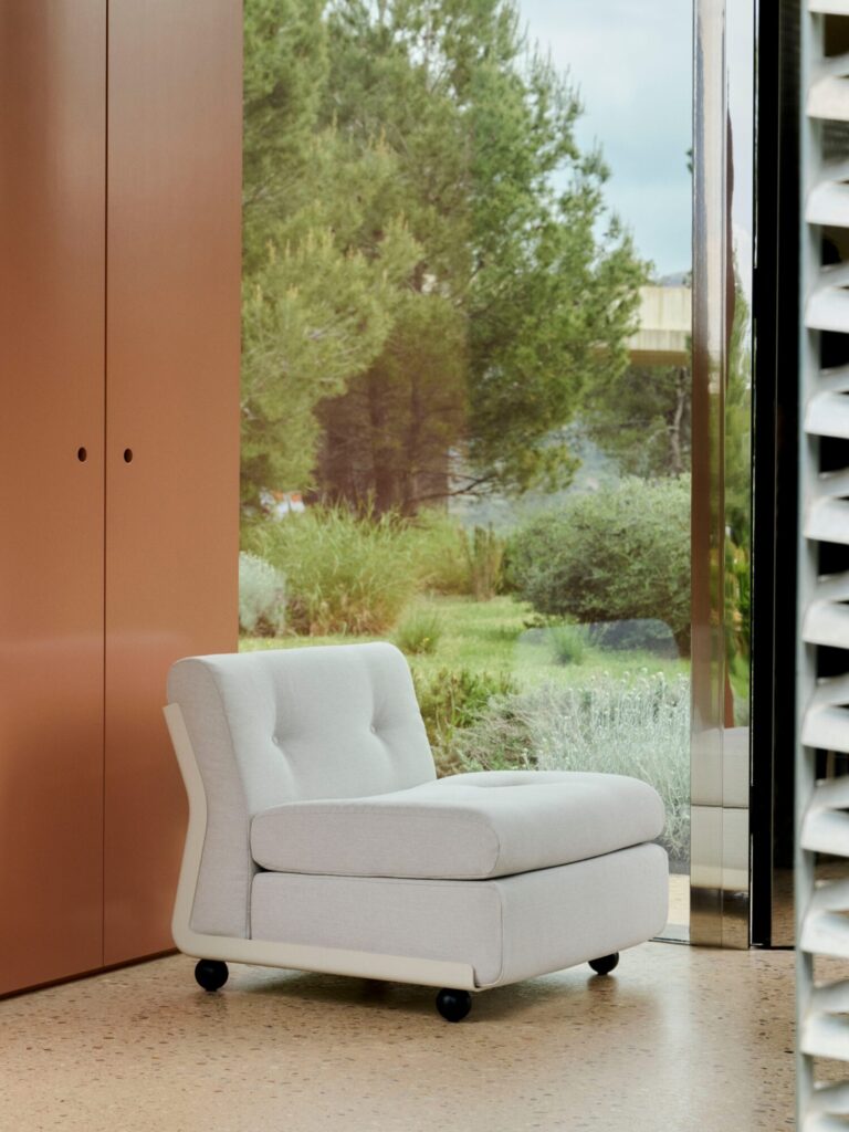 Un fauteuil Amanta 1 place gris clair à roulettes est placé à l'intérieur près d'une fenêtre, offrant une vue sur un jardin luxuriant avec des arbres et des buissons.