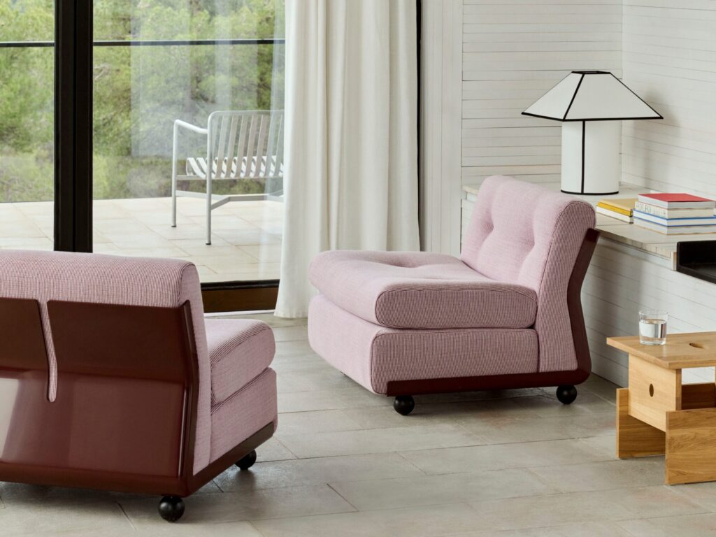 Le salon moderne comprend deux chaises roses Amanta à une place, une table d'appoint en bois avec une lampe et des livres, et une porte vitrée ouvrant sur un patio avec des sièges d'extérieur.