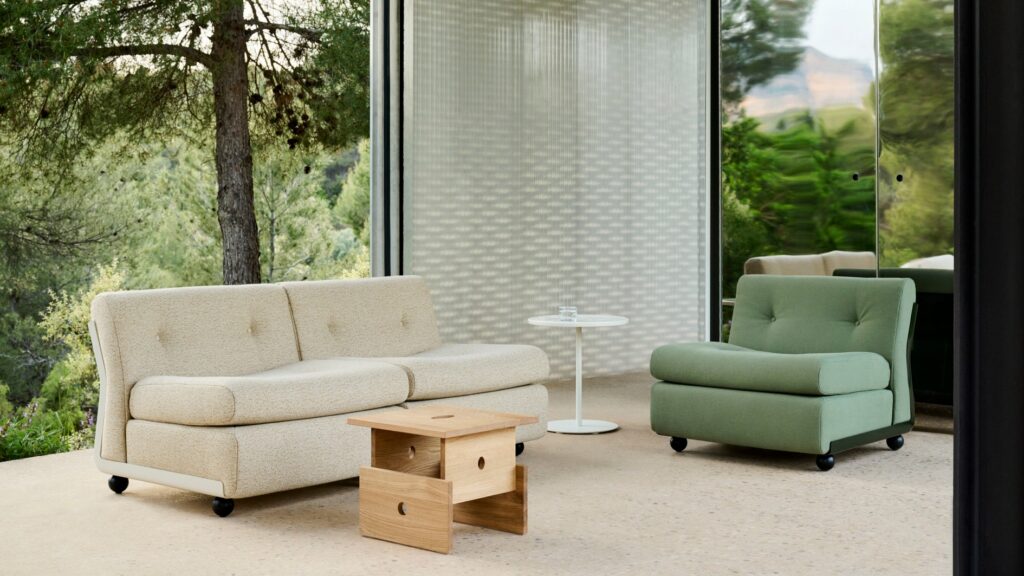 Un Amanta 2 places beige, un fauteuil vert, une petite table ronde blanche et une table d'appoint en bois sont disposés sur un patio couvert avec de grandes parois vitrées donnant sur les arbres.