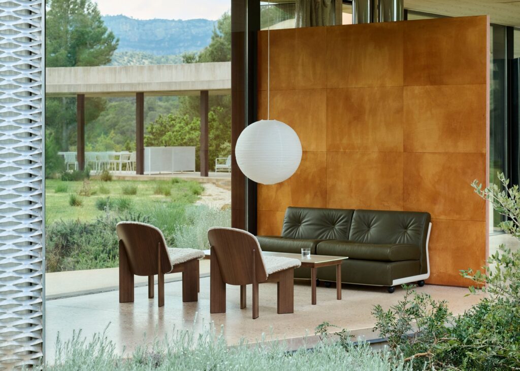 Salon moderne avec un fauteuil Amanta 2 places en vert foncé, deux chaises en bois, une petite table et une lampe suspendue ronde près de grandes fenêtres donnant sur un jardin et un patio minimalistes.