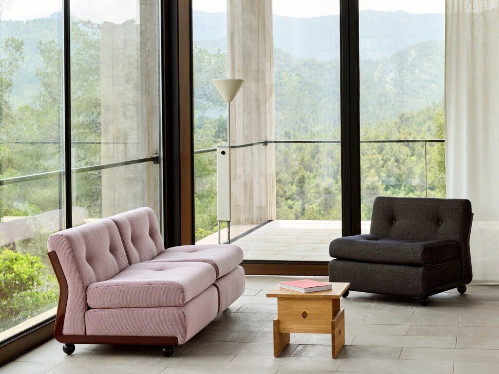 Un salon moderne avec un canapé Amanta 2 places rose, un fauteuil gris foncé, une table d'appoint en bois avec des livres, de grandes fenêtres et des vues sur les arbres et les montagnes.