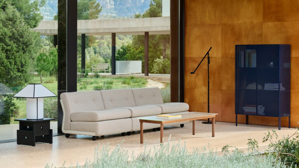 Salon moderne avec un canapé Amanta 3 places en Tissu groupe 1, une table basse en bois, un lampadaire noir, une armoire bleue et une lampe blanche sur une table d'appoint noire, le tout placé devant de grandes fenêtres donnant sur de la verdure.