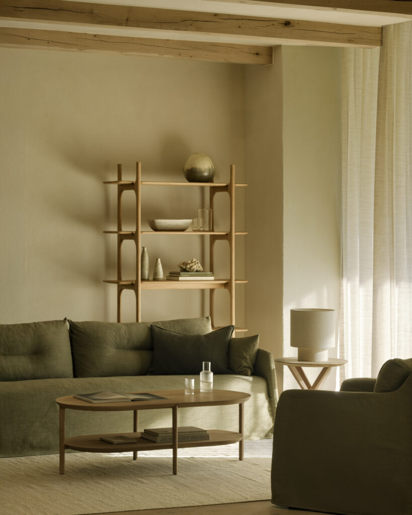 Un salon aux tons neutres avec un élégant canapé vert 3pl, une table basse en bois, des étagères ouvertes avec des objets décoratifs et des rideaux transparents qui laissent entrer la lumière naturelle.