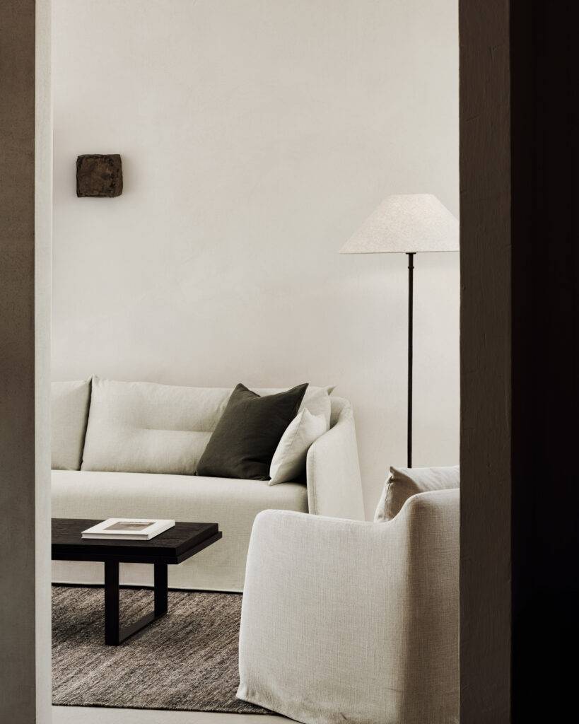 Salon minimaliste avec un canapé en tissu vert, une table basse noire contenant un livre, un lampadaire et un tapis marron contre un mur uni de couleur claire.