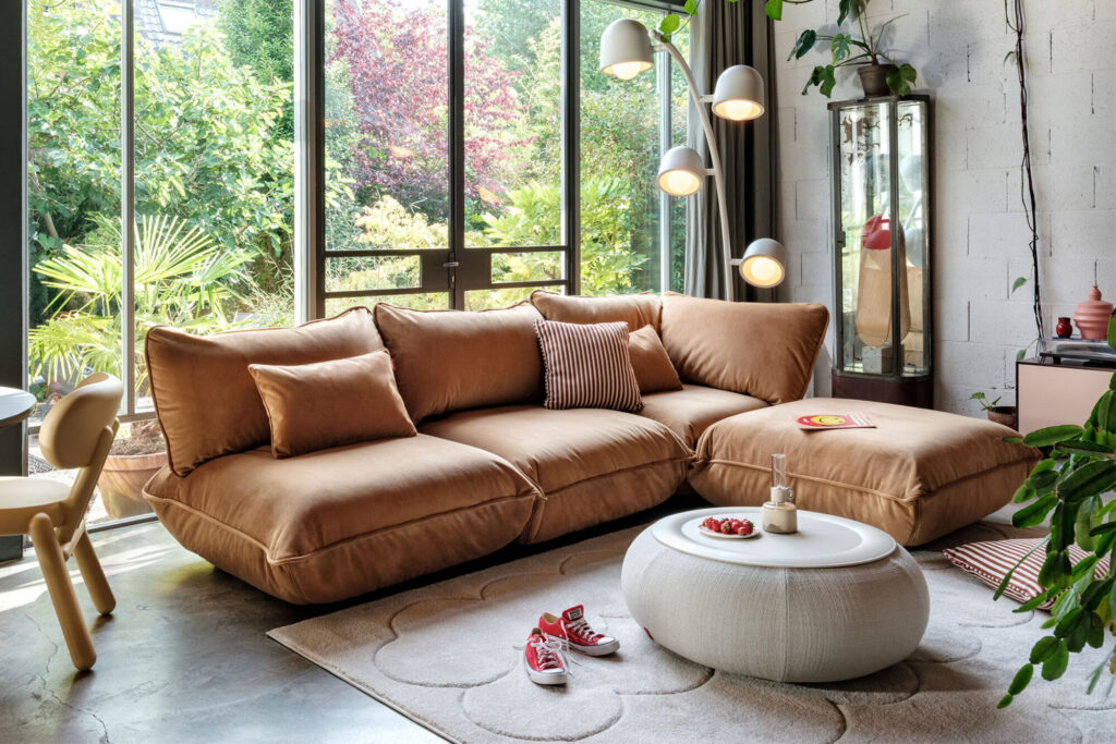 Un salon moderne avec un canapé sectionnel beige et un siège d'angle Sumo en velours recyclé vert foncé, un ottoman rond blanc, un lampadaire, des portes vitrées donnant sur un jardin et des plantes d'intérieur. Des baskets rouges et un livre sont posés sur le tapis.