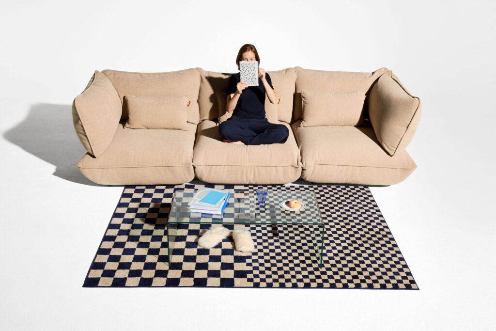 Une personne est assise les jambes croisées sur un siège d'angle Sumo beige, tenant un livre devant son visage, avec une table basse en verre placée sur un tapis à carreaux noir et blanc devant elle.