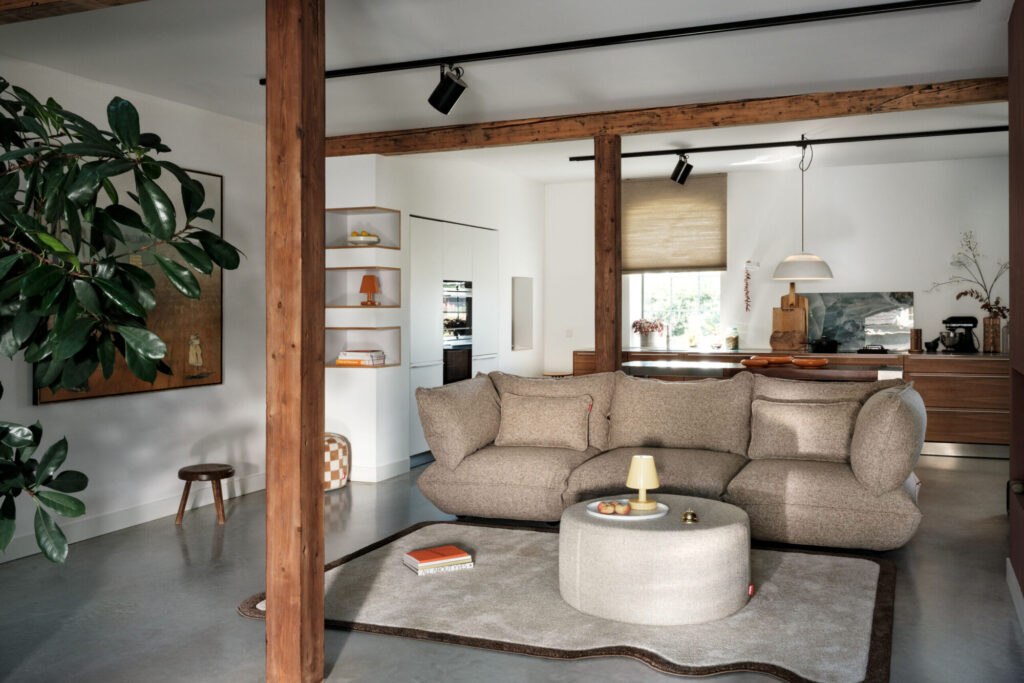 Salon moderne avec un canapé Bouclé beige, un ottoman rond, des livres, des poutres en bois, une grande plante et une cuisine ouverte en arrière-plan. La lumière naturelle pénètre par une fenêtre avec un abat-jour beige pour une touche à la fois confortable et recyclée.