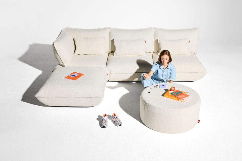 Une femme en tenue bleu clair est assise sur un ottoman rond près d'un siège d'angle Sumo beige recyclé avec texture bouclée, entourée de livres, de chaussures et de petits objets éparpillés sur un fond blanc.