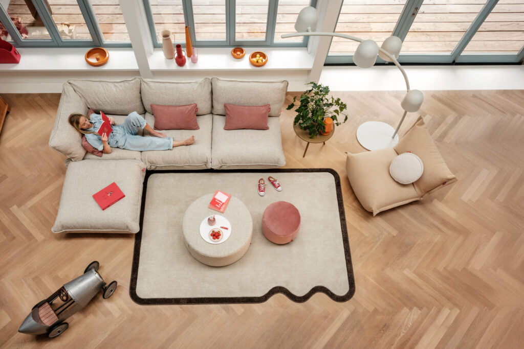 Un enfant est allongé sur un siège d'angle Sumo Beige recyclé et lit un livre dans un salon moderne avec des planchers en bois, une grande fenêtre et des meubles en bouclette contemporains.