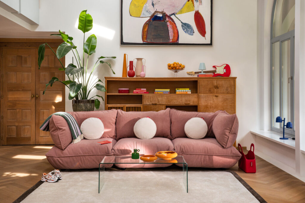 Un siège Sumo rose avec des coussins blancs ronds se trouve devant une étagère en bois avec une décoration colorée, une grande peinture abstraite, une table basse en verre et une grande plante dans ce salon lumineux.