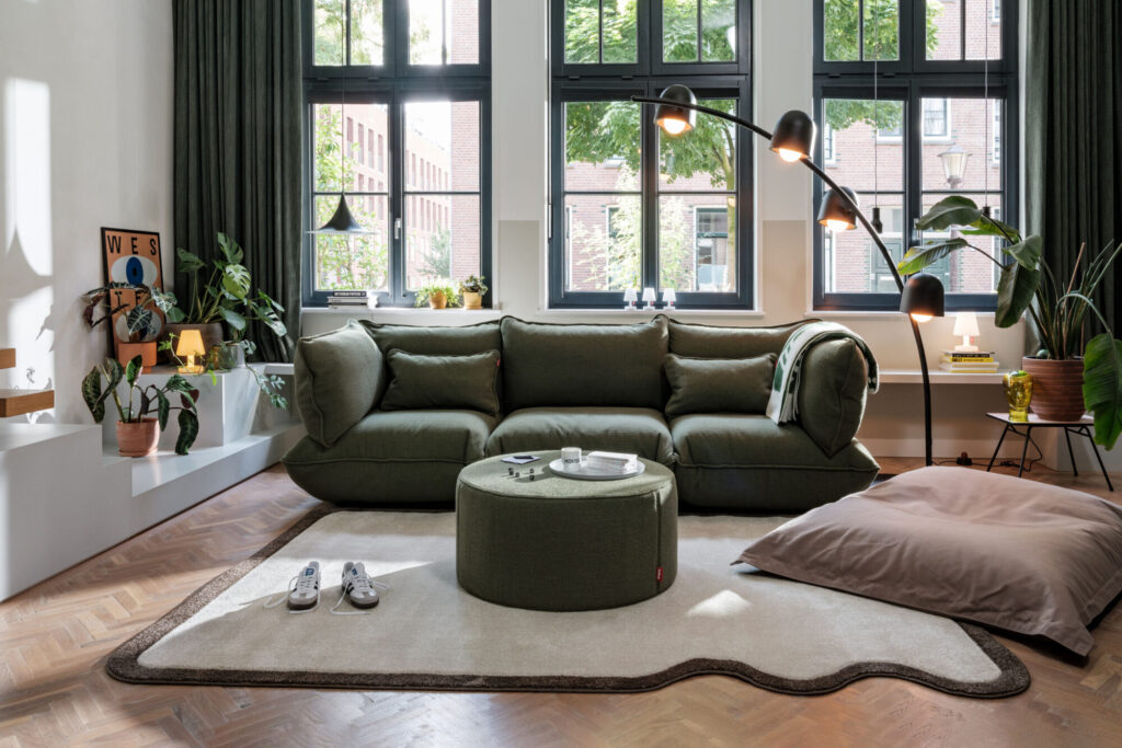 Un salon moderne avec un canapé en bouclette recyclé vert feuille, un ottoman assorti, un grand coussin de sol Sumo, des plantes en pot et un lampadaire, le tout entouré de hautes fenêtres aux cadres noirs et aux rideaux verts.