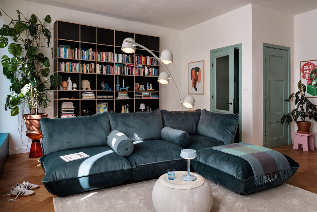 Salon moderne avec un siège Sumo recyclé en velours vert foncé, un lampadaire, des étagères, des plantes en pot et des œuvres d'art sur les murs.