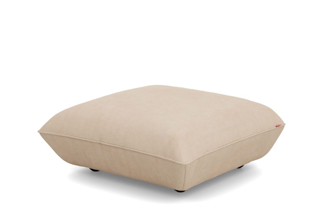 Un ottoman Sumo Hocker carré, en tissu beige, avec des bords arrondis et de courts pieds noirs, placé sur un fond blanc.