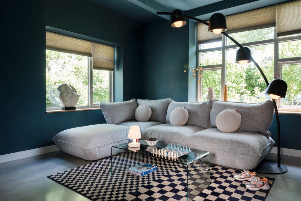 Salon moderne avec des murs sarcelle, un grand canapé sectionnel gris associé à un Sumo Hocker recyclé en Bouclé mélangé marron clair, un lampadaire arc, une table basse en verre avec un jeu d'échecs, un tapis à carreaux et de grandes fenêtres donnant sur de la verdure.