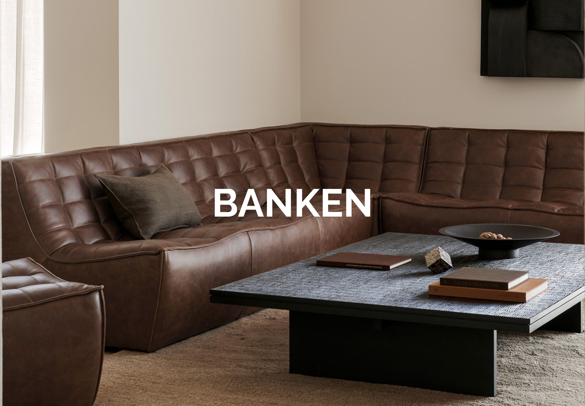Un salon moderne avec un canapé sectionnel en cuir marron, une table basse sombre avec des livres et un bol, le tout dans un style quatuor. Le mot "BANKEN" est superposé à l'image, évoquant l'ambiance chic d'un magasin de meubles.