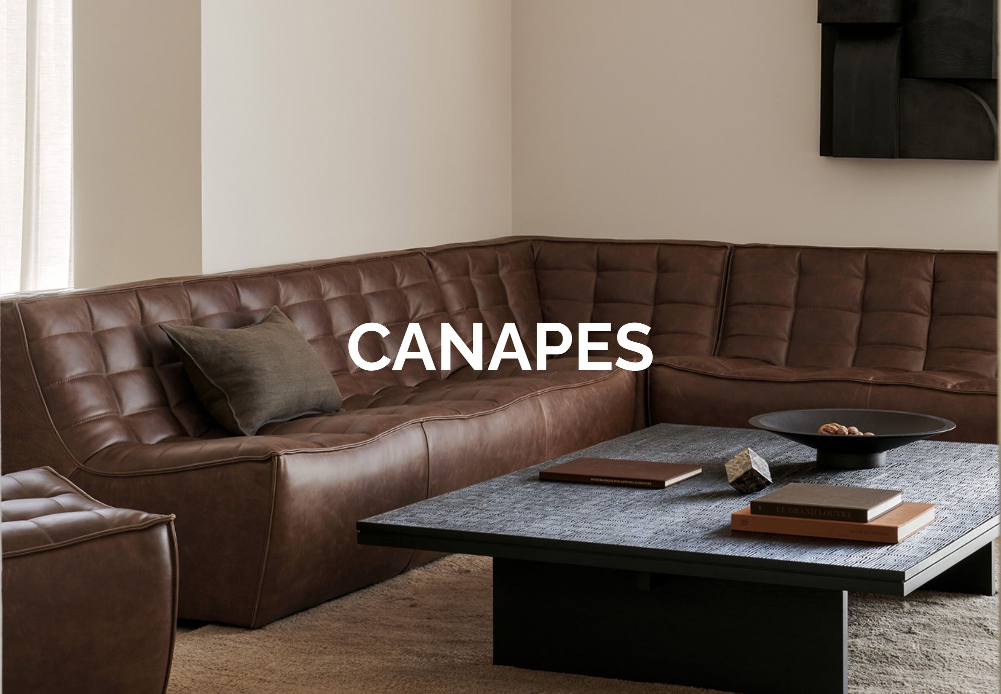 Un salon moderne est composé d'un canapé sectionnel en cuir marron, d'une table basse foncée avec des objets de décoration et de murs neutres. Le mot "CANAPES" est superposé en grandes lettres blanches, évoquant l'élégance du design quatuor que l'on retrouve dans tout magasin de meubles.