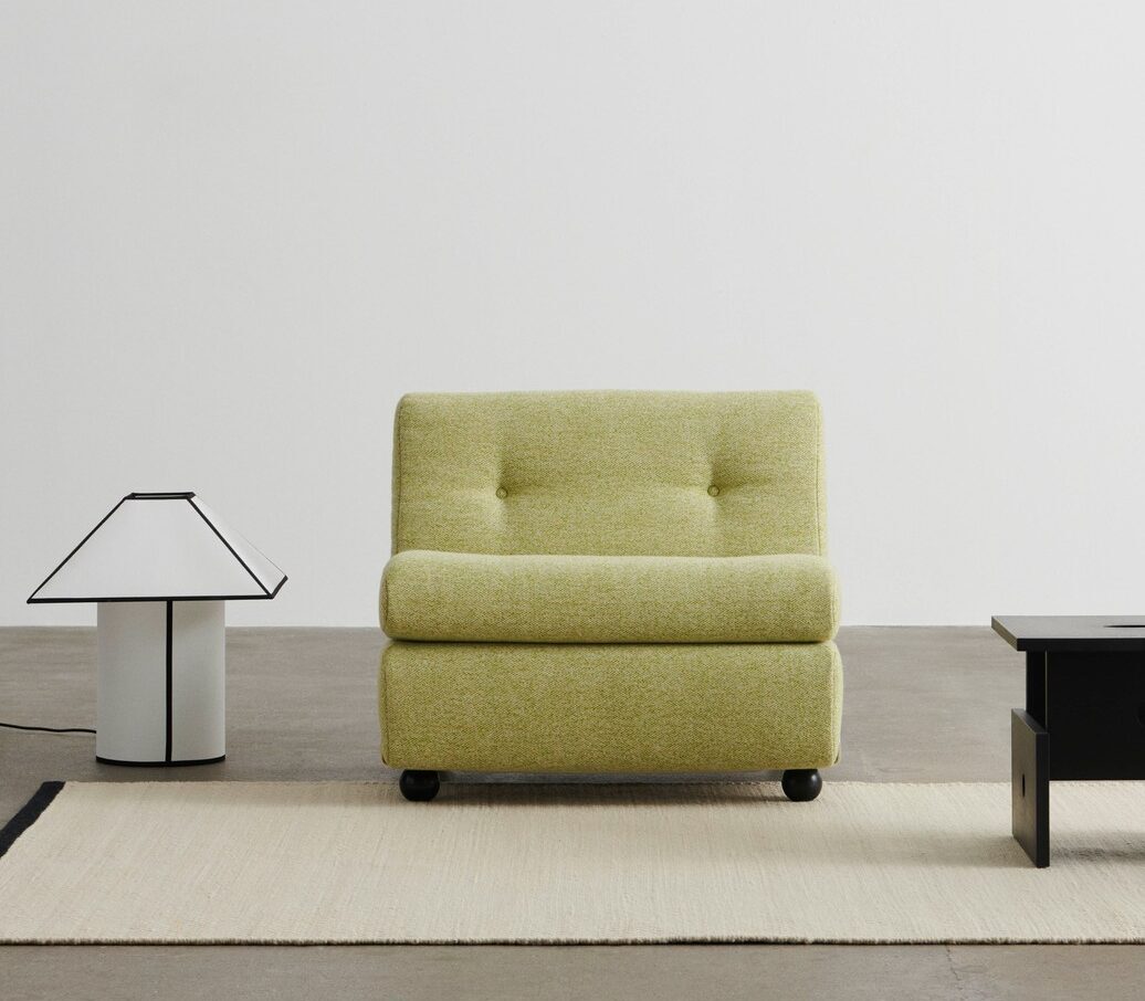 Un fauteuil vert sans accoudoir est posé sur un tapis beige entre une lampe de table blanche avec une garniture noire et une table basse noire, créant ainsi une ambiance October Deco Days sur un mur blanc uni.